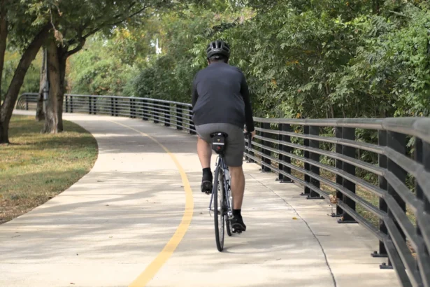 A man cyclist rides away on a paved, tree-lined trail with a black railing